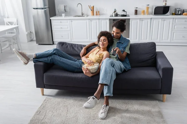 Sonriente afroamericano hombre sosteniendo mando a distancia cerca de novia con palomitas de maíz en casa - foto de stock