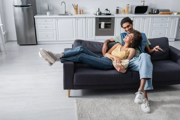 Cheerful african american couple holding bowl of popcorn and remote controller on couch — Foto stock