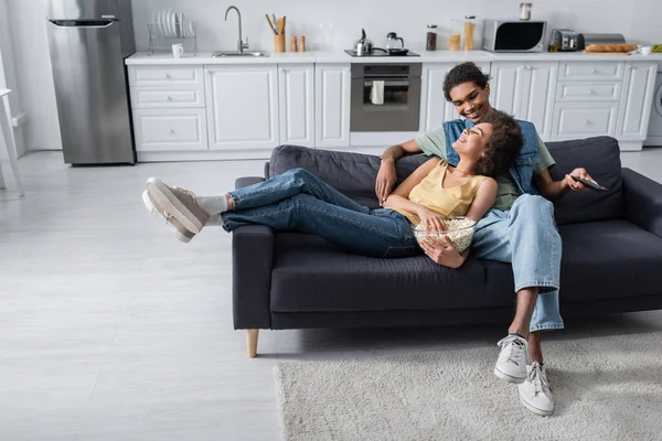 Cheerful african american couple holding bowl of popcorn and remote controller on couch — Stockfoto