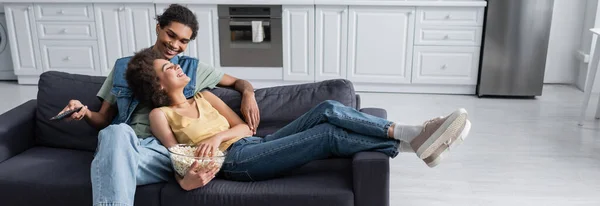 Felice coppia afro-americana che tiene popcorn e telecomando sul divano a casa, banner — Foto stock
