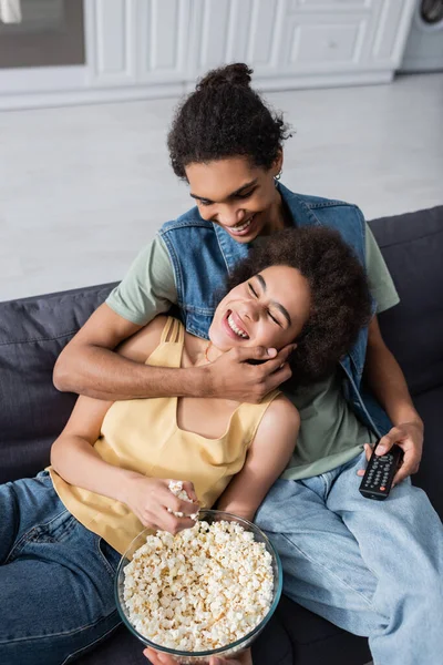 Vista de ángulo alto del hombre afroamericano sonriente sosteniendo el mando a distancia y tocando a la novia con palomitas de maíz en el sofá - foto de stock
