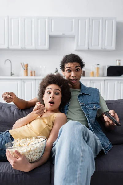 Casal afro-americano chocado segurando controlador remoto e pipoca no sofá em casa — Fotografia de Stock