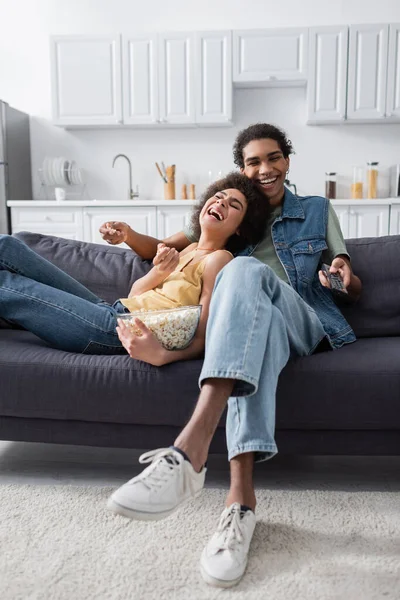 Riendo pareja afroamericana sosteniendo palomitas de maíz y controlador remoto en el sofá - foto de stock