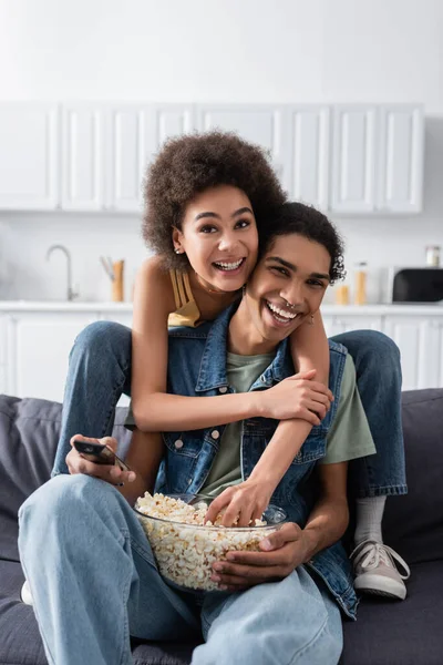 African american woman hugging smiling boyfriend with popcorn and remote controller on couch — Fotografia de Stock