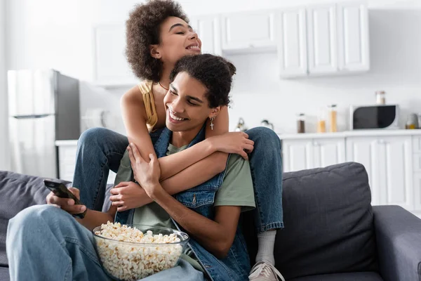 Donna afroamericana sorridente che abbraccia il fidanzato con telecomando e popcorn a casa — Foto stock