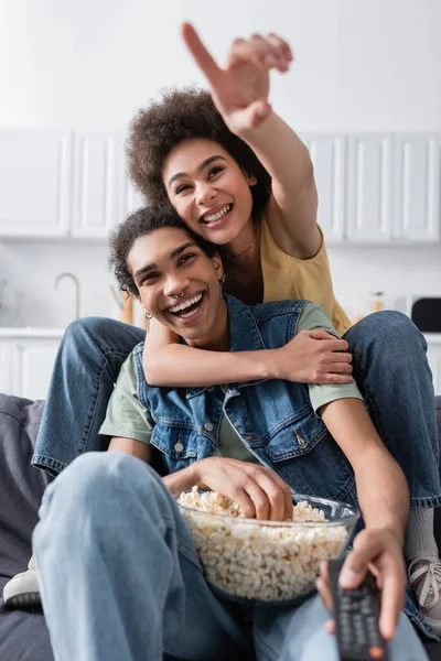 Mujer afroamericana señalando con el dedo y abrazando novio con mando a distancia y palomitas de maíz en casa - foto de stock