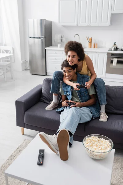 KYIV, UCRAINA - 9 NOVEMBRE 2021: Giovane coppia afroamericana che gioca ai videogiochi vicino ai popcorn e al telecomando a casa — Foto stock