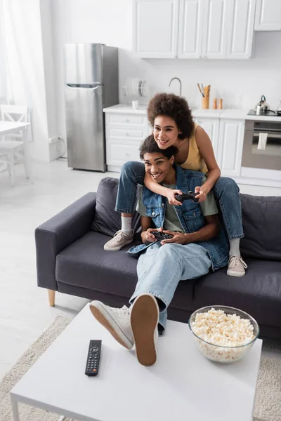 KYIV, UKRAINE - 9 NOVEMBRE 2021 : Jeune couple afro-américain jouant à un jeu vidéo près de popcorn et télécommande à la maison — Photo de stock