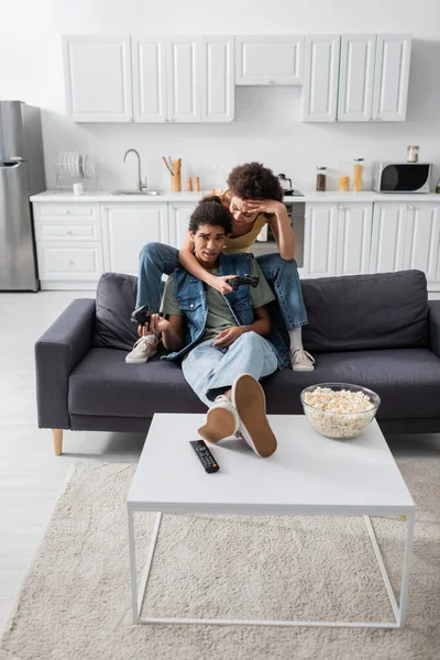 KYIV, UKRAINE - NOVEMBER 9, 2021: African american woman holding joystick near upset boyfriend and popcorn at home — Stockfoto