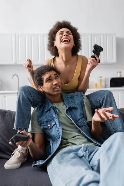 QUIIV, UCRÂNIA - NOVEMBRO 9, 2021: Mulher afro-americana entusiasmada segurando joystick perto namorado descontente no sofá — Fotografia de Stock