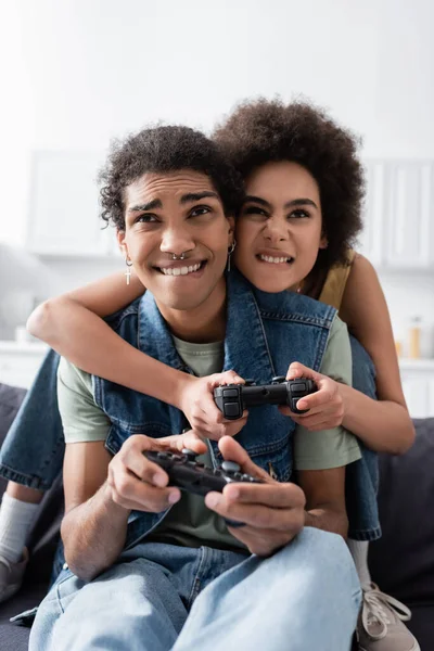 KYIV, UCRANIA - 9 de noviembre de 2021: Pareja afroamericana enfocada jugando videojuegos en el sofá en casa - foto de stock