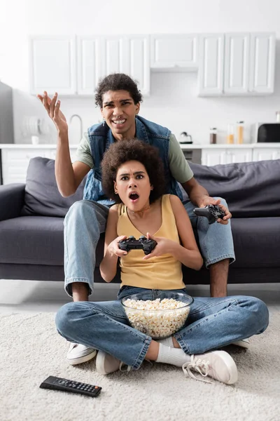 KYIV, UKRAINE - NOVEMBER 9, 2021:Dissapointed african american man holding joystick near shocked girlfriend and popcorn at home — Stock Photo