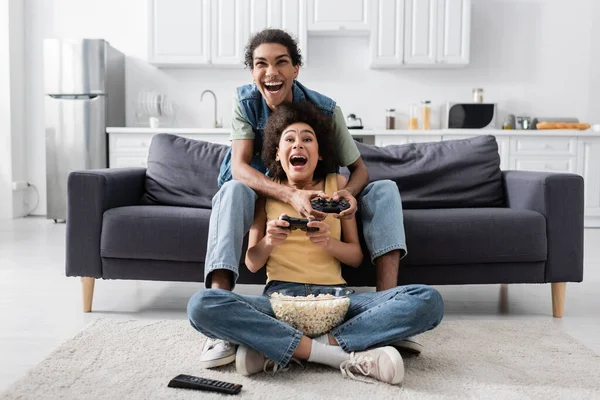 KIEW, UKRAINE - 9. NOVEMBER 2021: Aufgeregtes afrikanisch-amerikanisches Paar mit Popcorn beim Videospiel zu Hause — Stockfoto