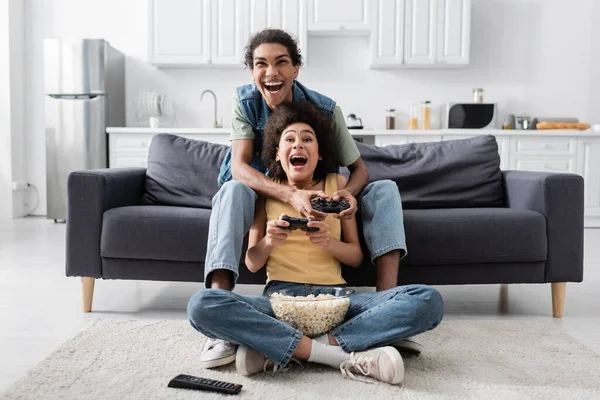 KYIV, UCRÂNIA - NOVEMBRO 9, 2021: Entusiasmado casal afro-americano com pipocas jogando videogame em casa — Fotografia de Stock