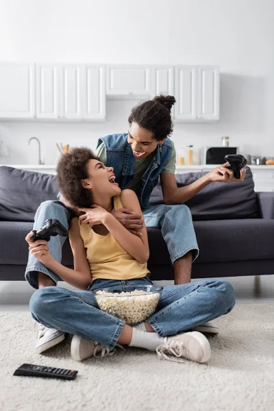 KYIV, UKRAINE - NOVEMBER 9, 2021: Excited african american man holding joystick and hugging girlfriend with popcorn at home — Stock Photo