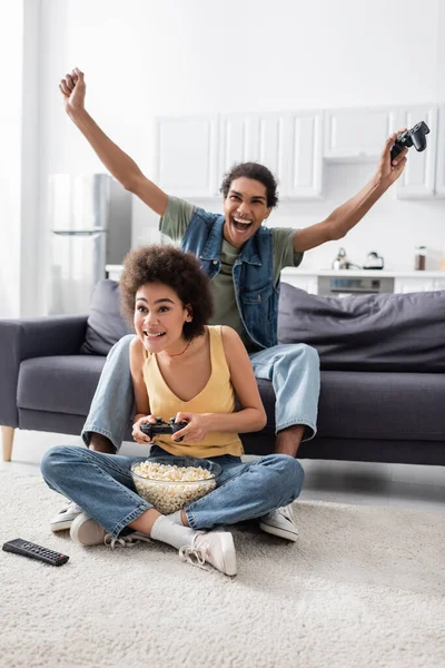 QUIIV, UCRÂNIA - NOVEMBRO 9, 2021: Emocionado afro-americano segurando joystick perto da namorada e pipocas em casa — Fotografia de Stock