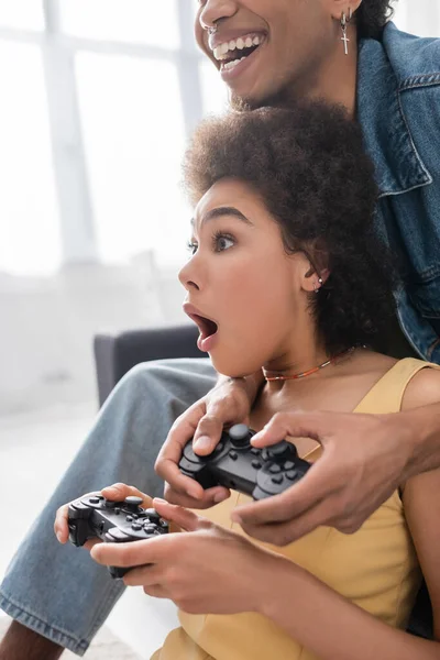 KYIV, UKRAINE - NOVEMBER 9, 2021: Shocked african american woman playing video game with smiling boyfriend at home — Stock Photo