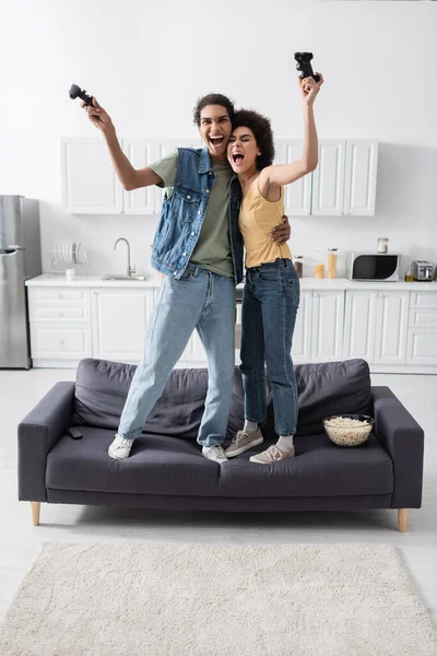 KYIV, UKRAINE - NOVEMBER 9, 2021: Excited african american couple holding joysticks on couch near popcorn and remote controller — Stock Photo