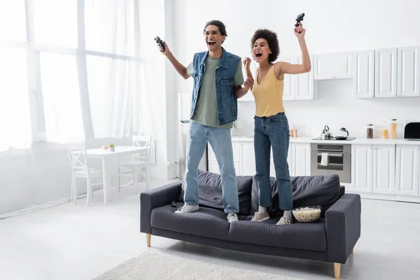 KYIV, UKRAINE - NOVEMBER 9, 2021: Excited african american couple with joysticks standing on couch near popcorn and remote controller — Stock Photo