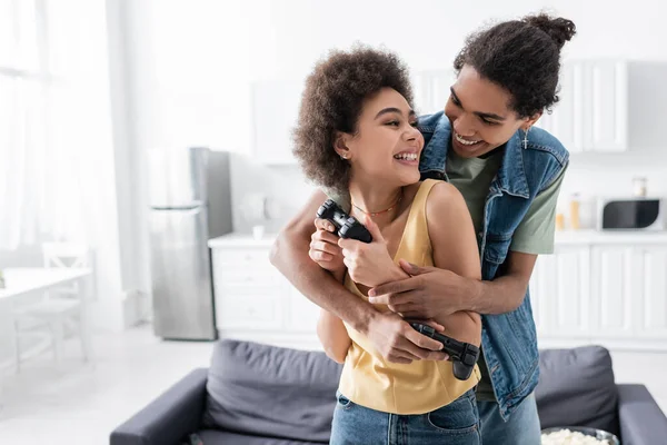 KYIV, UKRAINE - NOVEMBER 9, 2021: Smiling african american man embracing girlfriend with joystick at home — Fotografia de Stock