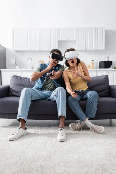 KYIV, UCRANIA - 9 de noviembre de 2021: Pareja afroamericana conmocionada en auriculares vr con joysticks en el sofá - foto de stock