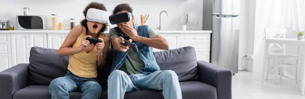 KYIV, UCRAINA - 9 NOVEMBRE 2021: Coppia afroamericana scioccata che gioca ai videogiochi e usa cuffie vr a casa, banner — Foto stock