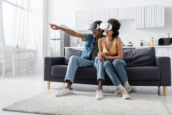 Hombre afroamericano en auriculares vr apuntando con el dedo cerca de la novia en el sofá - foto de stock