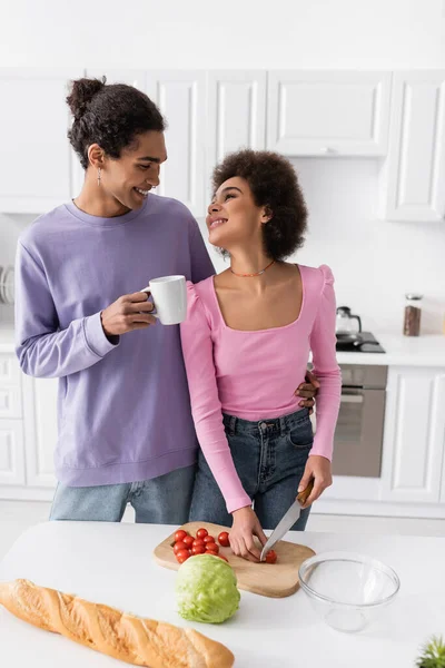 Smiling man holding cup and hugging african amerian girlfriend cutting cherry tomato at home — Stockfoto