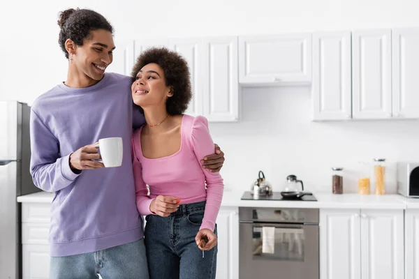 Souriant homme afro-américain avec tasse étreignant petite amie avec tomate cerise et couteau à la maison — Photo de stock