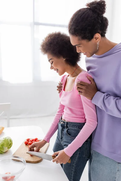 Vista lateral de un joven afroamericano abrazando a su novia cortando ensalada en casa - foto de stock