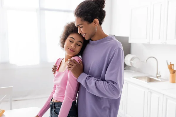 Alegre afroamericano hombre abrazando novia con los ojos cerrados en casa - foto de stock