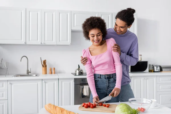 Jeune homme afro-américain étreignant petite amie couper des tomates cerises près de tasse et baguette à la maison — Photo de stock