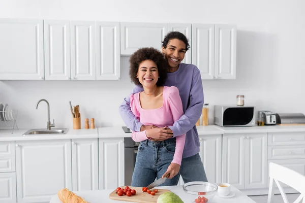 Sonriente afroamericano hombre abrazando novia cerca de verduras y café en casa - foto de stock