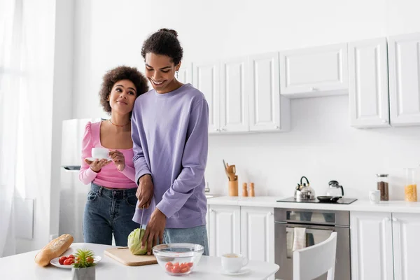 Young african american woman holding cup near boyfriend cutting cabbage and baguette at home — Stock Photo