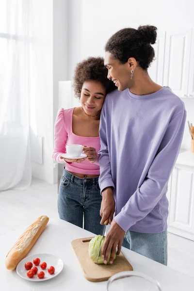 Souriant homme afro-américain coupe chou près de petite amie avec café dans la cuisine — Photo de stock