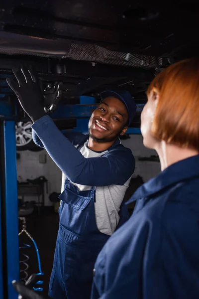 Sonriente afroamericano mecánico mirando borrosa colega cerca de auto en servicio - foto de stock