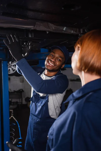 Sorridente meccanico afroamericano guardando collega offuscata vicino auto in servizio — Foto stock