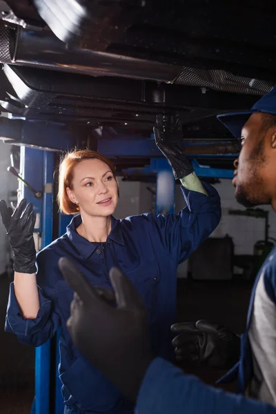 Smiling mechanic in uniform looking at african american colleague near car in service — стоковое фото