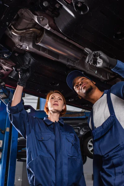 Low angle view of multiethnic mechanics pointing at car bottom in service — стоковое фото