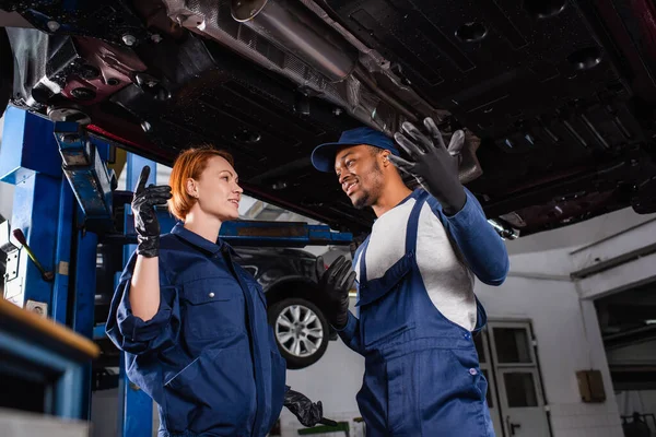 Mecánica interracial positiva en guantes y uniforme de pie bajo el coche en servicio - foto de stock