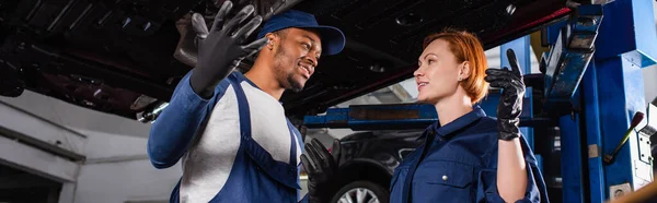 Mecánica interracial sonriente apuntando a la parte inferior del coche en el servicio automático, pancarta - foto de stock