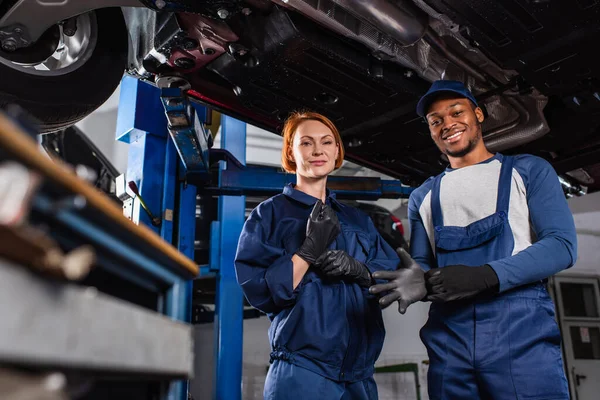 Cheerful multiethnic mechanics wearing gloves under car in service — Stock Photo