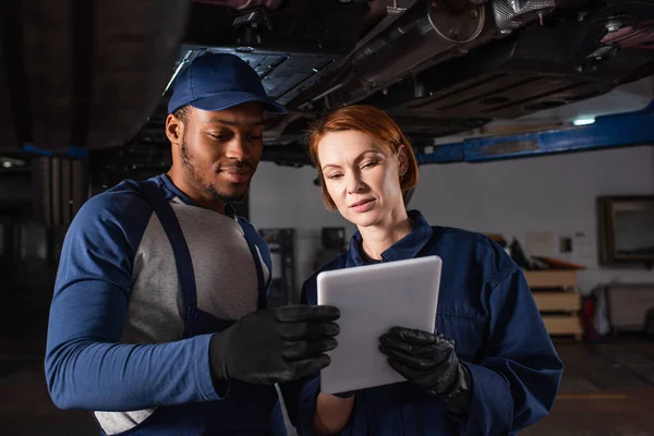 Multiethnic mechanics in gloves using digital tablet under car in garage — Stock Photo