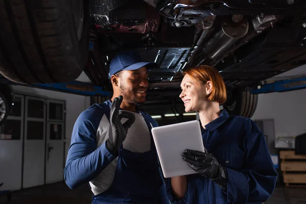 Smiling african american mechanic showing ok gesture near colleague with digital tablet under car in service — Stockfoto