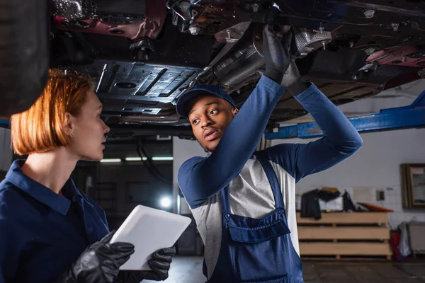 Trabajadora sosteniendo tableta digital cerca de un colega afroamericano debajo del coche en el garaje - foto de stock