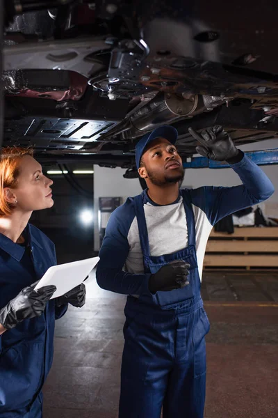 African american mechanic in uniform pointing at car bottom near colleague with digital tablet in service — Stock Photo