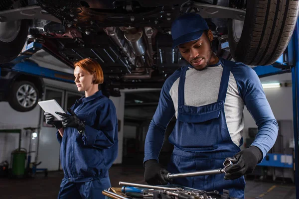 African american mechanic in gloves holding wrench near tools and colleague with digital tablet in car service — Fotografia de Stock