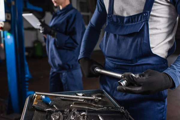Ausgeschnittene Ansicht eines Mechanikers, der Schraubenschlüssel neben verschwommenem Kollegen mit digitalem Tablet in der Garage hält — Stockfoto
