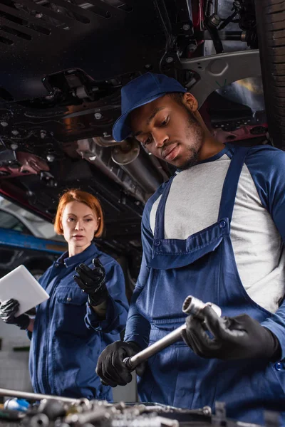 Afrikanischer Mechaniker hält Schraubenschlüssel neben verschwommenem Kollegen mit digitalem Tablet und Auto in Garage — Stockfoto