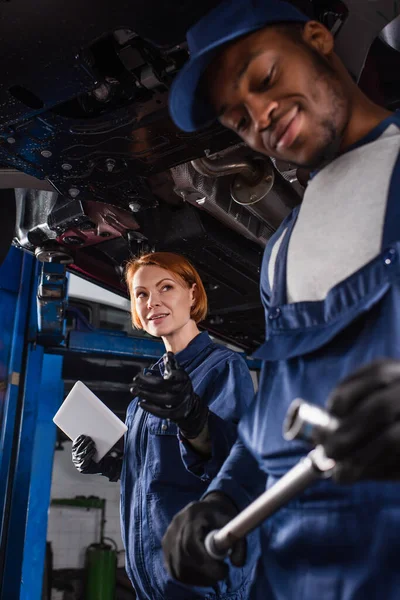 Low angle view of smiling workwoman holding digital tablet and pointing at african american colleague with wrench in car service — Stockfoto